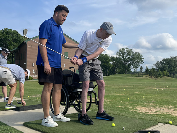 A participant in the golf clinic is assisted with swinging at a golf ball.