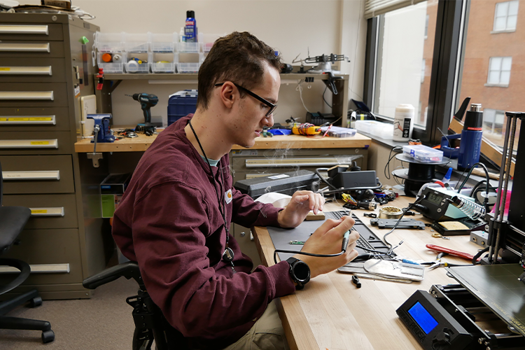 The Rehabilitation Engineer is seen handling a piece of electrical equipment as he actively engages in the creation of a personalized assistive technology device within his rehabilitation engineering lab.