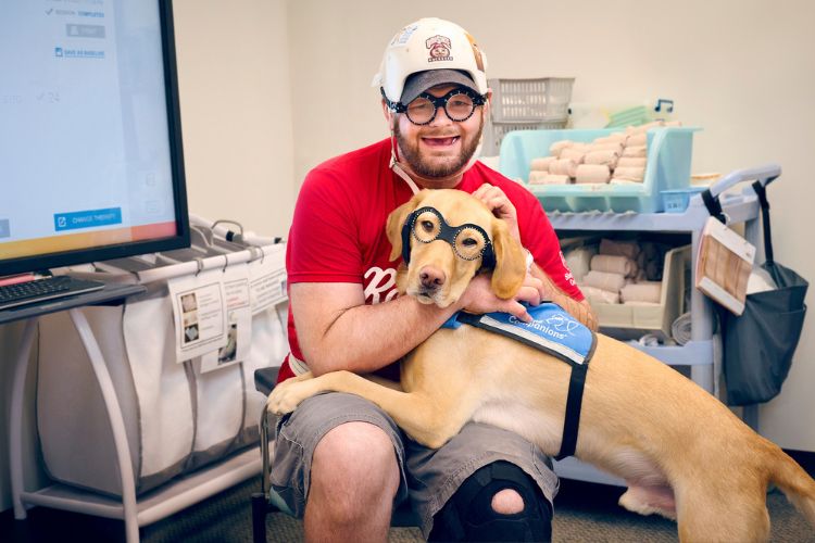 Patient smiles with Shepherd facility dog, Tex. They are both wearing prism glasses to help the visual midline to shift to a more centered position to expand visual awareness after brain injury. Tex is the official spokes pup of Shepherd's prism glasses awareness and education campaign, helping to normalize the use of prism glasses for patients with brain injuries.