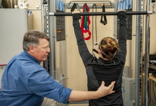 The physical therapist in the Pain Institute hold's a patients mid back as she reaches her arms high for the pull down bar during therapy