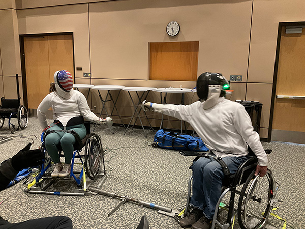 Two individuals in wheelchairs and full fencing gear practice their en garde stance