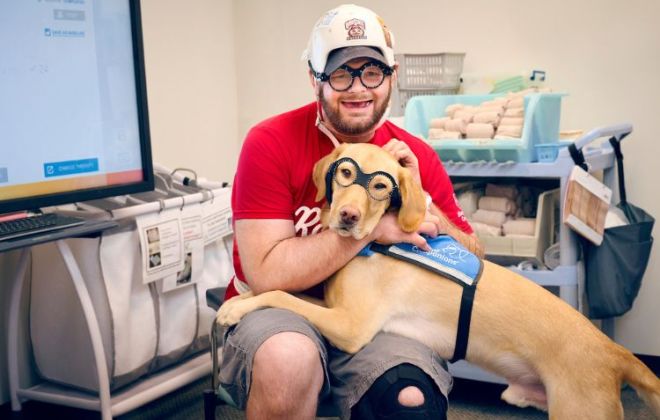 Patient wearing prism glasses to help with some visual symptoms of traumatic brain injury poses with facility dog, Tex, who also sports his own version of prism glasses.