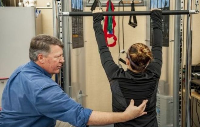The physical therapist in the Pain Institute hold's a patients mid back as she reaches her arms high for the pull down bar during therapy