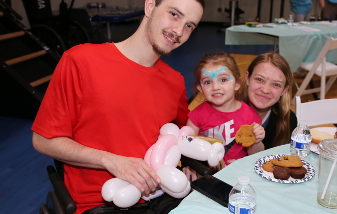 A spinal cord injury patient holds a balloon animal while smiling with his daughter and wife at a peer support event.
