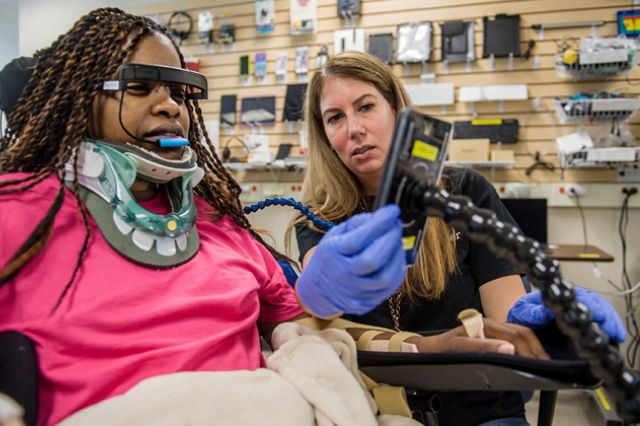Patient with a neck brace using a wheelchair and assistive technology tools in conjunction with speech therapy