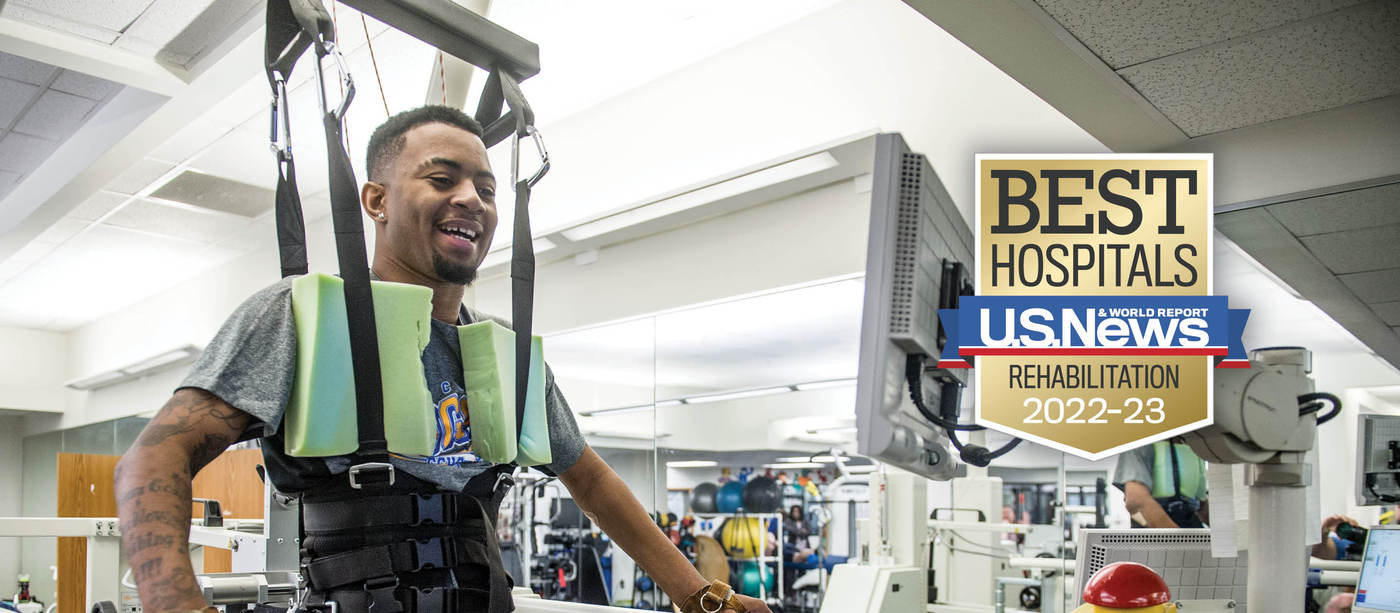 Shepherd Center patient smiles while using therapeutic equipment for lower-limb muscle strength. The U.S. News badge is overlayed with the words, "Best Hospitals Rehabilitation 2022-23."